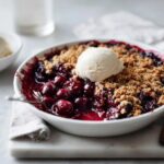 A warm bowl of Cherry Chocolate Crumble topped with a scoop of vanilla ice cream, served on a marble surface.