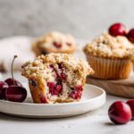 A close-up of a Cherry Oat Streusel Muffin cut in half showing the bright red cherry filling, next to fresh cherries.