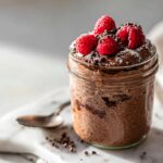 A jar of rich Chocolate Protein Overnight Oats topped with fresh raspberries and cacao nibs.