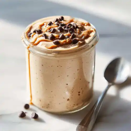 A jar of thick, blended Cookie Dough Blended Overnight Oats topped with peanut butter swirl and chocolate chips.