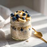A glass jar filled with creamy High Protein Overnight Oats, topped with sliced banana and fresh blueberries.