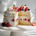 A slice removed from a rustic layer cake filled with cream and berries, decorated with fresh fruit and flowers for Rustic Wedding Dessert Table Ideas.