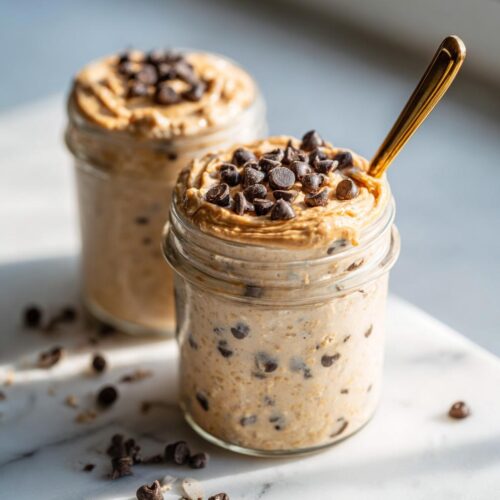 Two jars of Vegan Cookie Dough Overnight Oats topped with peanut butter swirl and chocolate chips, one with a gold spoon.