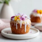 A close-up of one Wildflower Vanilla Cupcake topped with white glaze and fresh edible flowers.