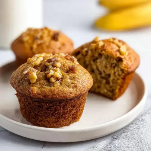Three freshly baked Banana Nut Muffins, one cut open, served on a white plate with walnuts on top.