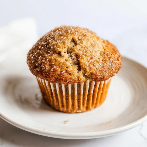 A single, perfectly domed Banana Muffins Bakery Style topped with coarse sugar, sitting on a white plate.