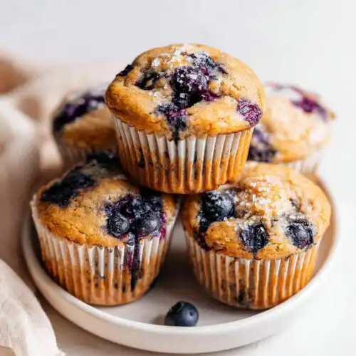 A stack of freshly baked Banana Blueberry Muffins topped with coarse sugar on a white plate.