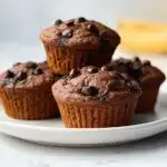 Four rich, dark Banana Chocolate Chip Muffins topped with melted chocolate chips, stacked on a white plate with a banana blurred in the background.