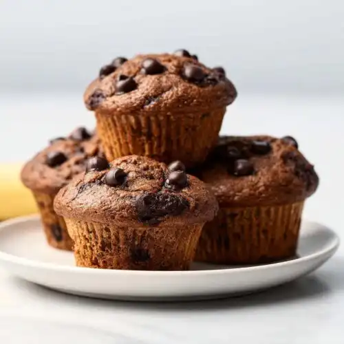 A close-up of four freshly baked Banana Chocolate Chip Muffins stacked on a white plate.