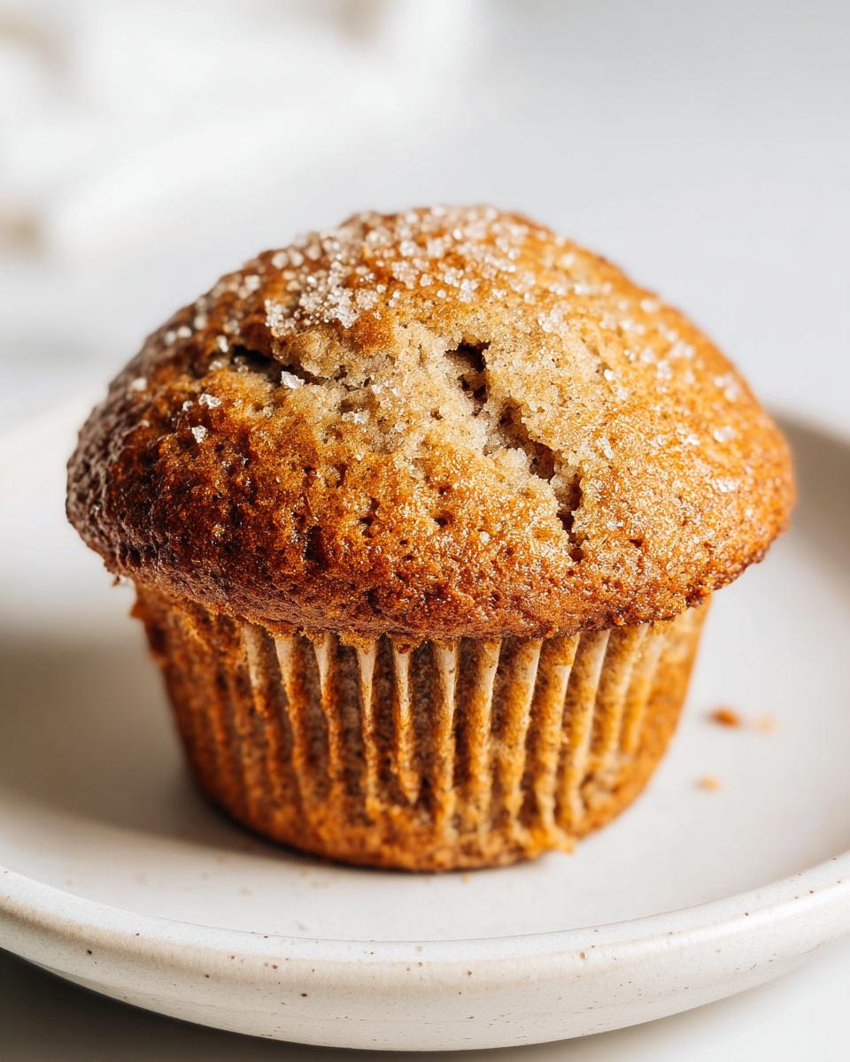 A single, perfectly domed Banana Muffins Bakery Style, topped with coarse sugar crystals, resting on a light ceramic plate.
