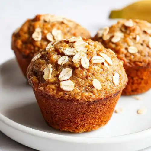 Close-up of three freshly baked Banana Oat Muffins topped with visible rolled oats on a white plate.