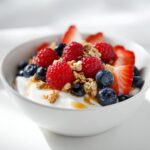 A close-up of a white bowl filled with yogurt, topped with fresh raspberries, blueberries, sliced strawberries, and granola for a Berry Breakfast Bowl.