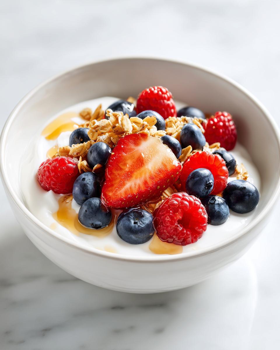 A close-up of a Berry Breakfast Bowl featuring yogurt, fresh blueberries, raspberries, sliced strawberry, granola, and a drizzle of honey.