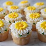 A close-up of several vanilla cupcakes topped with white and yellow buttercream daisy flowers, featuring yellow centers.