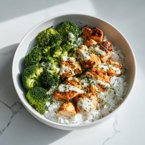 A white bowl containing rice, grilled chicken pieces, steamed broccoli, and a creamy herb sauce, perfect for High Protein Summer Meal Prep.