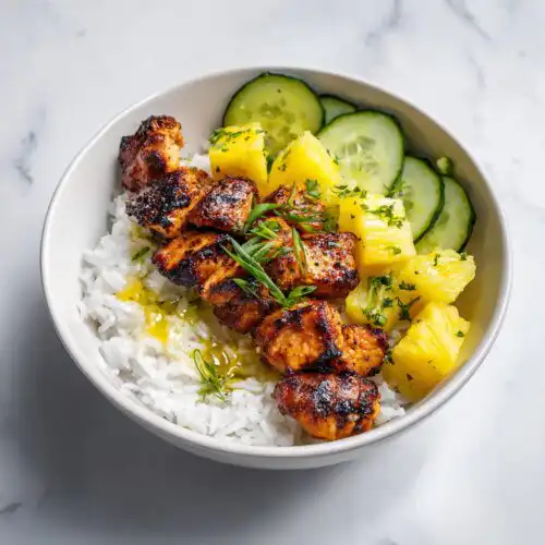 A white bowl filled with white rice, topped with grilled chicken pieces, pineapple chunks, and sliced cucumbers for a Chicken Pineapple Summer Bowl.