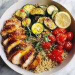 A vibrant bowl featuring sliced seasoned chicken breast over quinoa, served with grilled zucchini, roasted tomatoes, and a lemon slice, perfect for Light Summer Dinner Recipes.