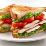 Two halves of a grilled Chicken Sandwich Picnic stacked high with lettuce, tomato, and sliced chicken breast on toasted bread.