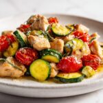 A close-up of the Chicken Summer Skillet Dinner featuring sautéed chicken pieces, zucchini rounds, and burst cherry tomatoes, topped with cheese.