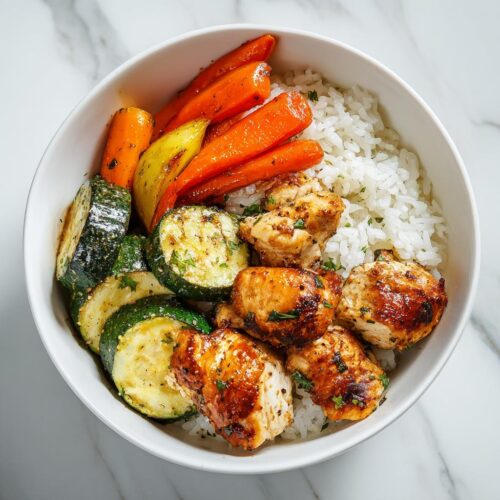 A close-up overhead view of a Chicken Veggie Summer Bowl featuring seasoned chicken pieces, grilled zucchini, carrots, and white rice.