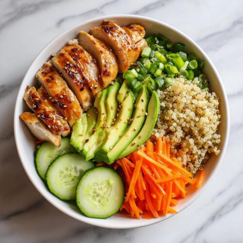 Overhead view of a vibrant Cold Chicken Bowl featuring sliced glazed chicken, avocado, quinoa, carrots, and cucumbers.