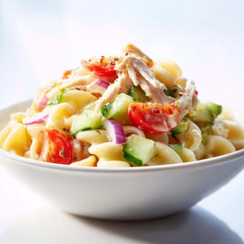 Close-up of Cold Chicken Pasta Salad served in a white bowl, featuring pasta, shredded chicken, cucumber, tomato, and red onion.