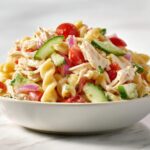 A generous serving of Cold Chicken Pasta Salad featuring rotini pasta, shredded chicken, cucumber slices, tomatoes, and red onion.