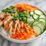 A close-up of a Cold Chicken Rice Bowl featuring sliced glazed chicken, white rice, shredded carrots, sliced cucumbers, and green onions.