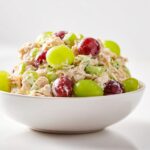 A white bowl filled with creamy Cold Chicken Salad Meal Prep mixed with celery and topped with red and green grapes.