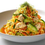 A beautifully plated Cold Noodle Chicken Bowl featuring noodles, shredded carrots, cucumber slices, and chicken pieces topped with sesame seeds.