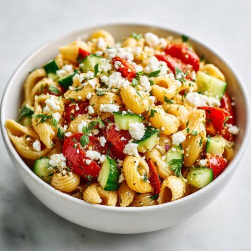 A close-up of a white bowl filled with Cold Pasta Salad Lunch featuring shell pasta, chopped cucumbers, tomatoes, and crumbled feta cheese.