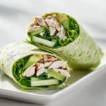 Two halves of a vibrant green tortilla filled with shredded chicken, fresh lettuce, cucumber, and avocado, making up a Cold Summer Chicken Wrap.