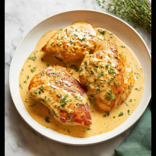 Three golden-brown chicken breasts swimming in a rich, creamy Dijon mustard sauce, garnished with fresh thyme.