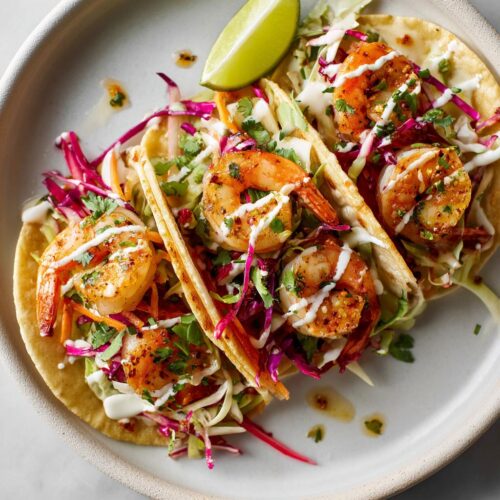 Three Easy Summer Shrimp Tacos filled with seasoned shrimp, colorful slaw, and drizzled with white sauce, served with a lime wedge.