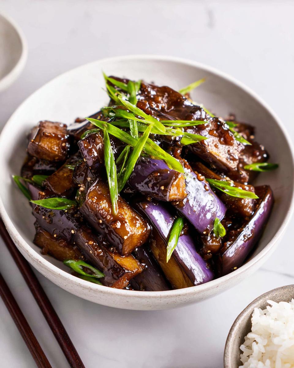 Close-up of glossy Eggplant Stir Fry Asian Style pieces topped with bright green scallions in a light ceramic bowl.