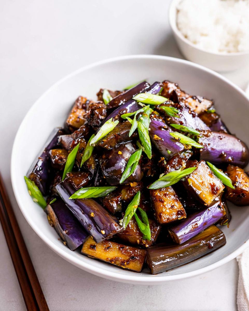 A close-up of glossy Eggplant Stir Fry Asian Style pieces topped with bright green scallions in a white bowl.