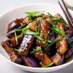Close-up of glossy Eggplant Stir Fry Asian Style pieces topped with green onions and sesame seeds in a white bowl.