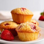 A stack of moist Gluten Free Strawberry Muffins topped with coarse sugar, served with fresh strawberries.