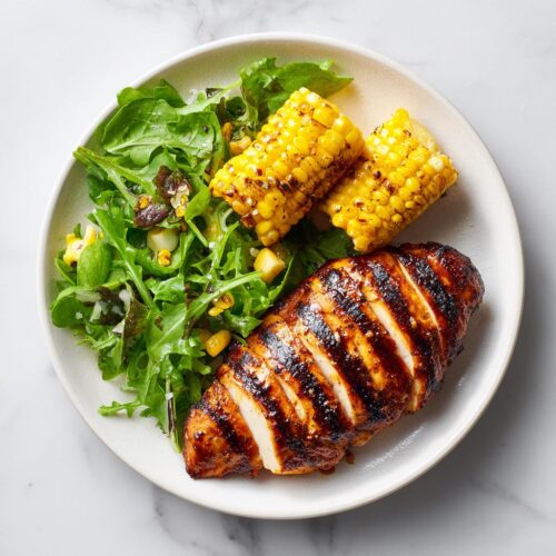 Overhead view of a perfectly sliced Grilled Chicken Summer Plate featuring BBQ chicken breast, grilled corn on the cob, and a fresh green salad.
