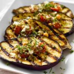 Four slices of perfectly Grilled Eggplant with Herbs, topped with quinoa, chickpeas, tomatoes, and feta.