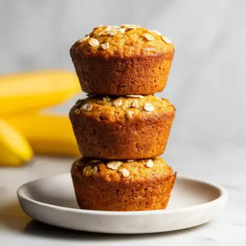Three golden brown Healthy Banana Breakfast Muffins stacked on a small white plate, topped with rolled oats.
