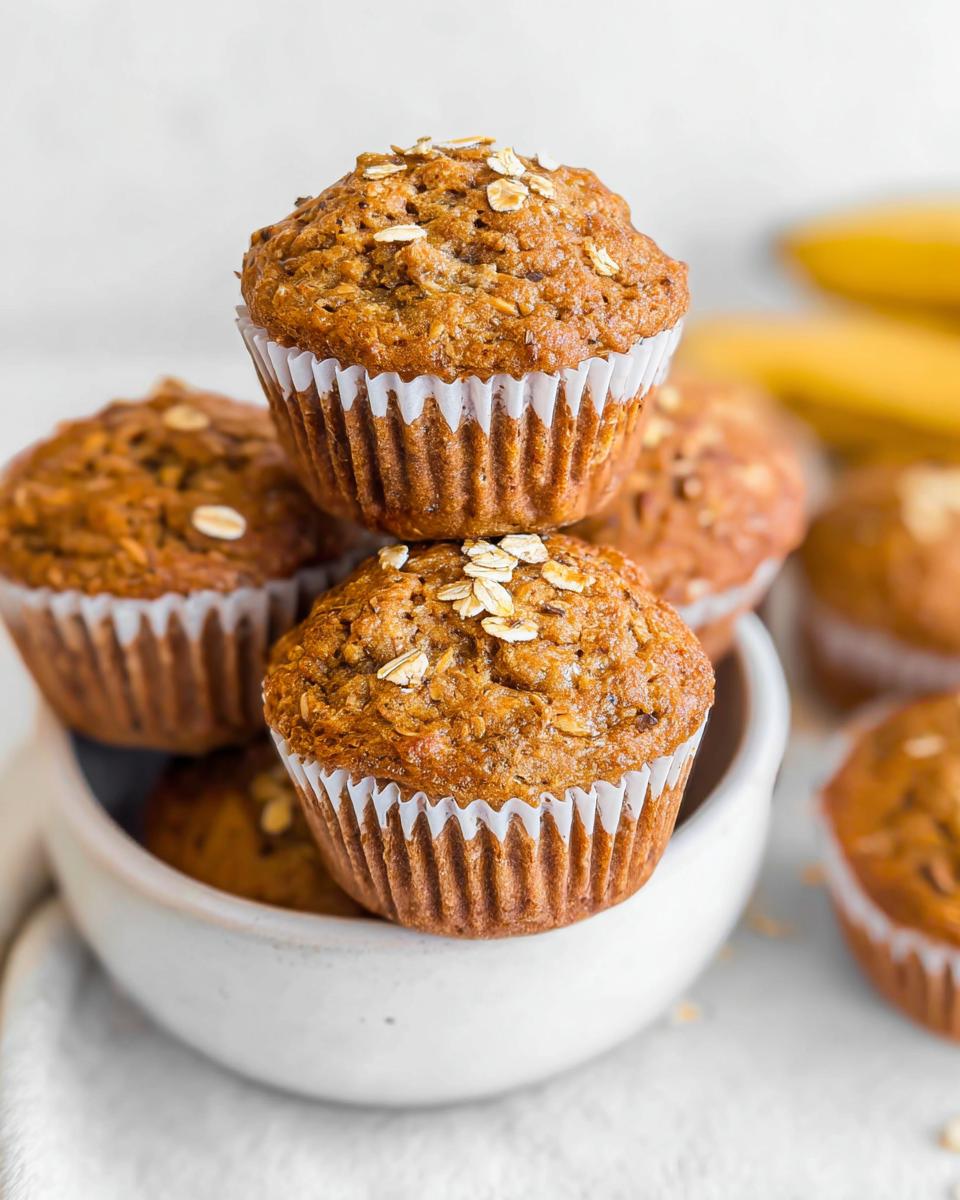 A stack of freshly baked Healthy Banana Muffins topped with rolled oats, sitting in a white bowl.