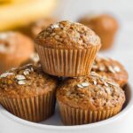 A close-up stack of four freshly baked Healthy Banana Muffins topped with rolled oats, resting in a white bowl.