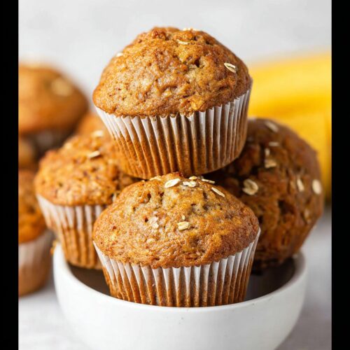 A stack of freshly baked Healthy Banana Muffins topped with rolled oats, presented in a white bowl.