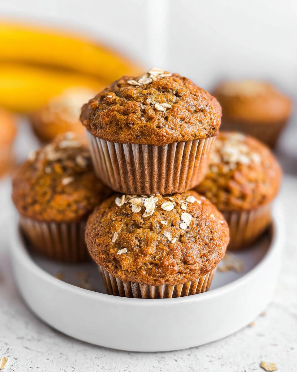 A stack of three freshly baked Healthy Banana Muffins topped with rolled oats, served in a white bowl.