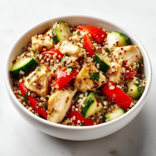 Close-up of a bowl containing Healthy Summer Chicken Lunch with quinoa, grilled chicken pieces, red bell peppers, and cucumber slices.
