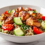 Close-up of a bowl containing a Healthy Summer Chicken Lunch with glazed chicken pieces over quinoa, cucumbers, and cherry tomatoes.
