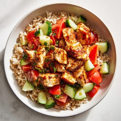Overhead view of a bowl containing brown rice topped with seasoned chicken pieces, diced tomatoes, and cucumber slices, perfect for Healthy Summer Lunch Ideas.