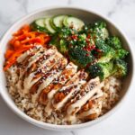 A vibrant Healthy Summer Protein Bowl featuring sliced grilled chicken over brown rice, topped with creamy sauce, sesame seeds, broccoli, carrots, and cucumber.