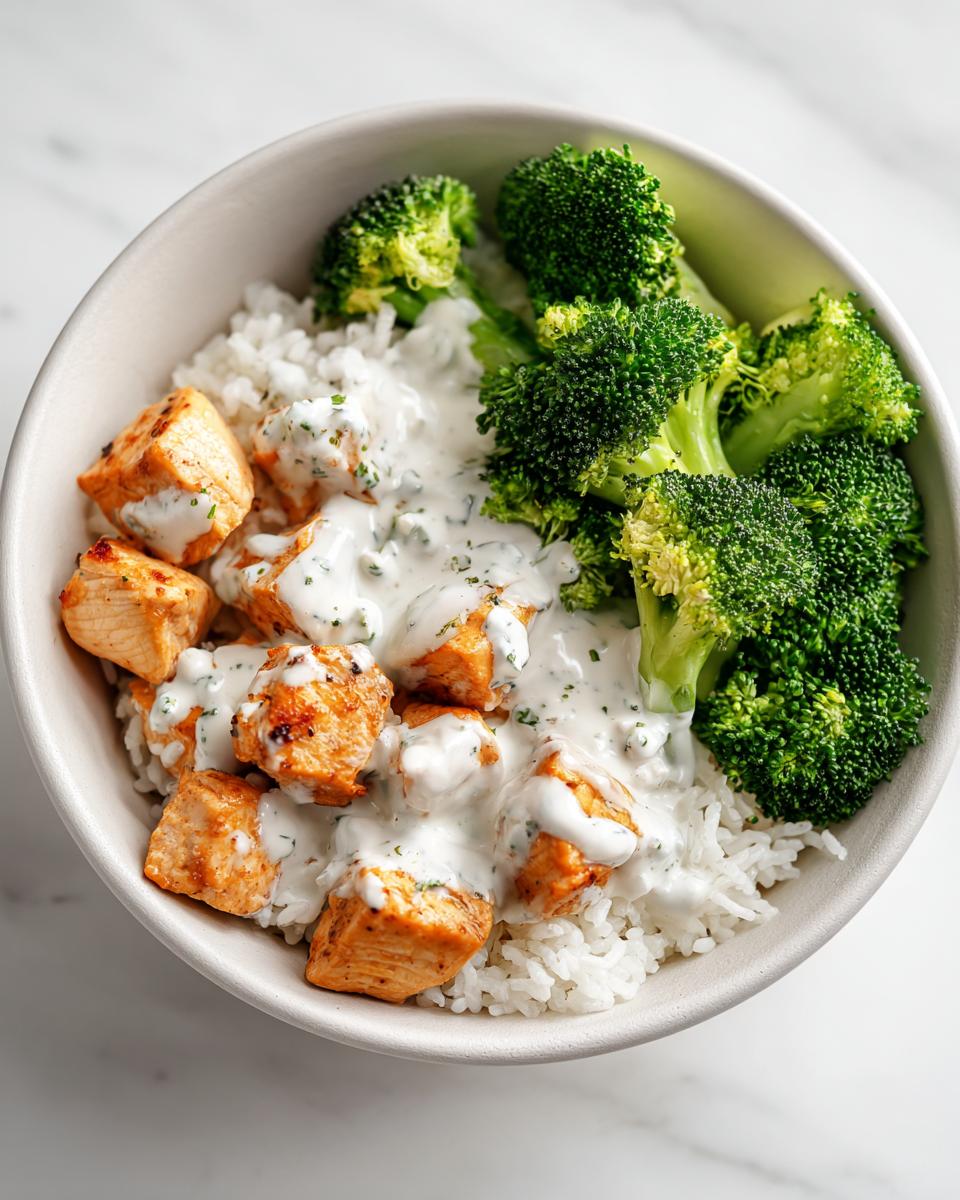 Close-up of a High Protein Chicken Bowl Meal Prep featuring seasoned chicken cubes, white rice, steamed broccoli, and a creamy herb sauce.